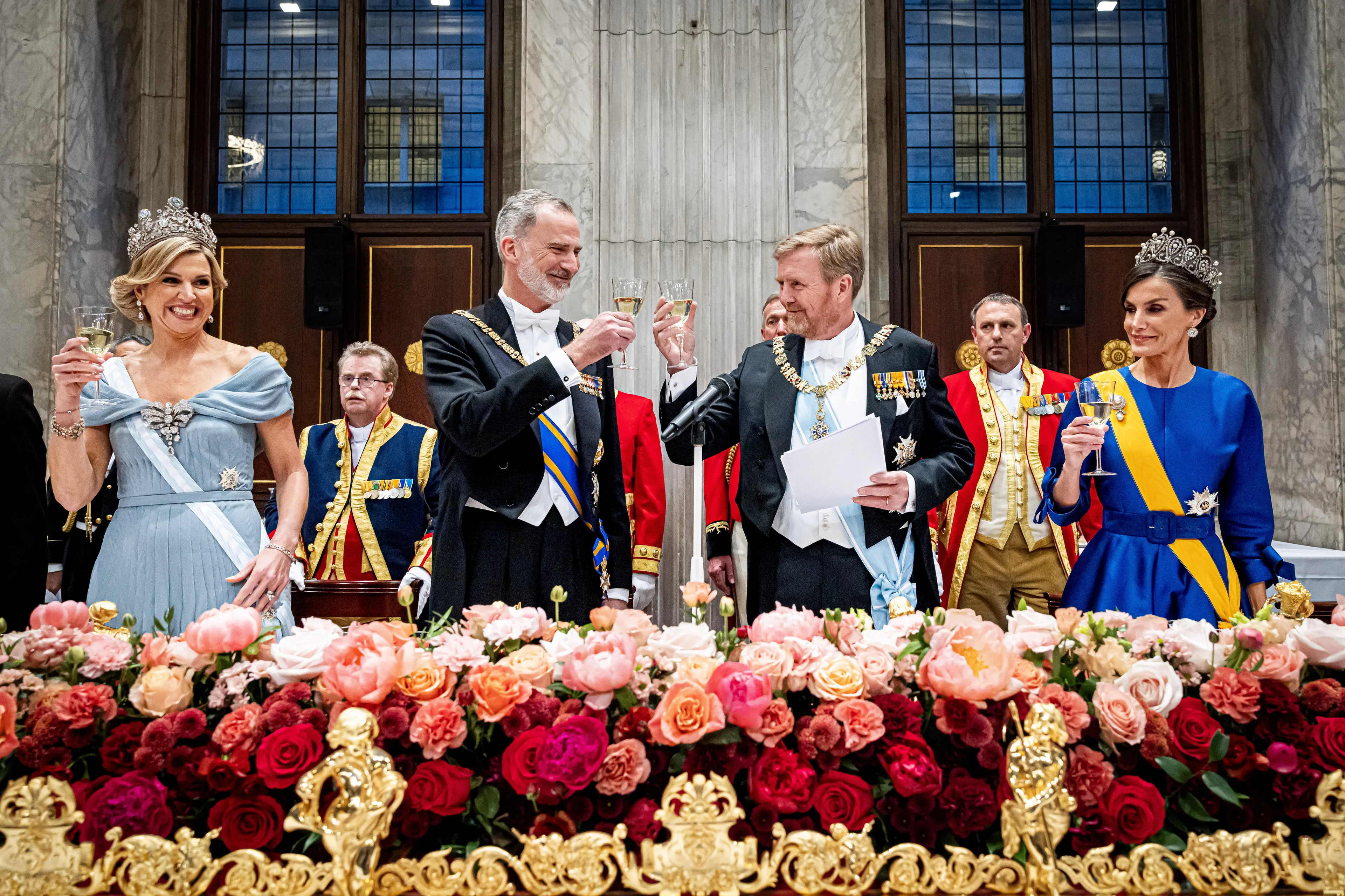 Staatsbanket of receptie: Een formele zaal met een lange, rijk versierde tafel waar twee koninklijke paren en de koning van Zweden aanwezig zijn, terwijl Koning Carl XVI Gustaf bij een microfoon een toost lijkt uit te spreken en het gezelschap het moment observeert.