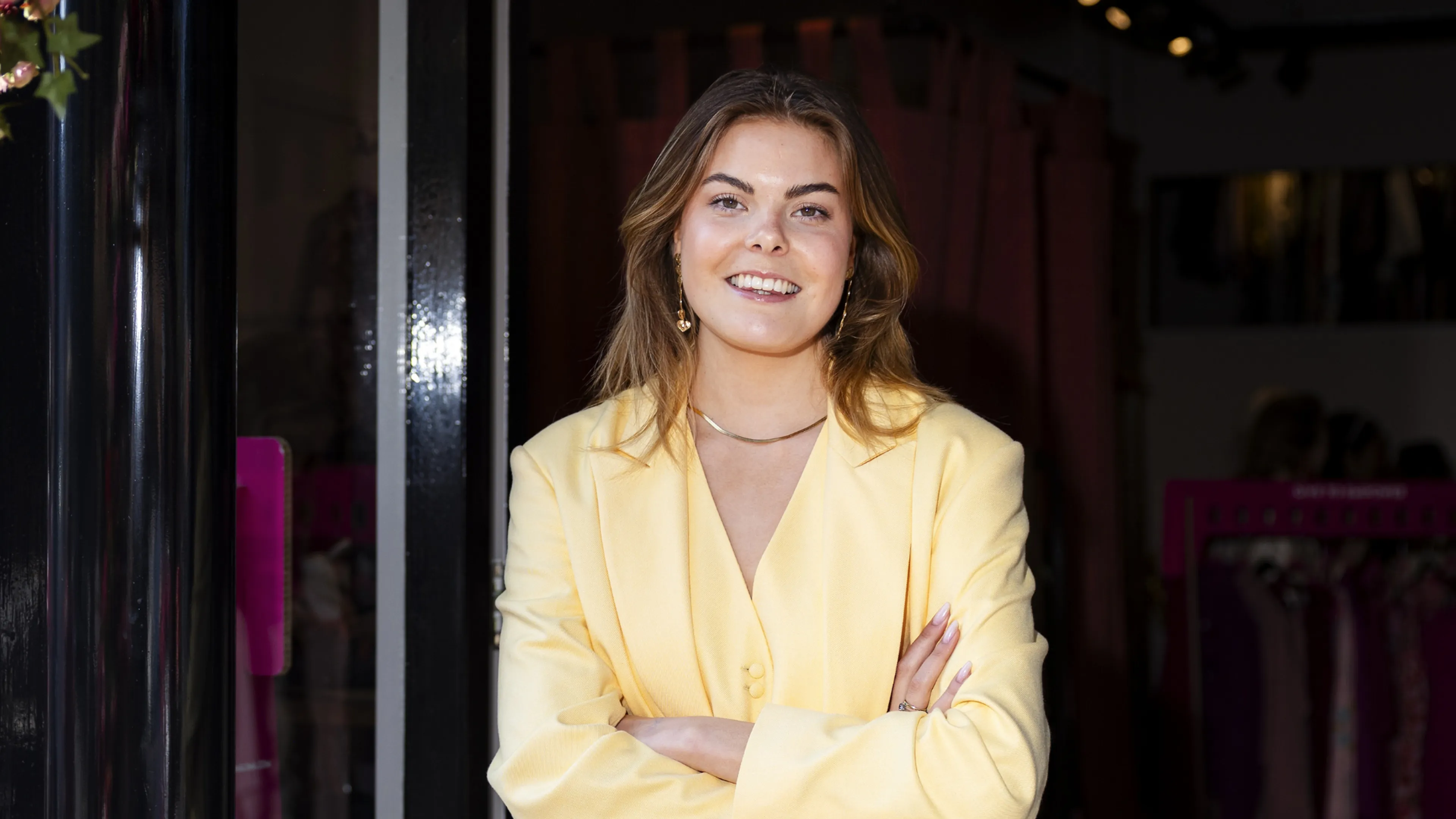 Beroemd geen specifieke naam genoemd. Een jonge vrouw staat centraal bij de ingang van een kledingboetiek, kijkend naar de camera met een open glimlach terwijl ze haar armen ontspannen over elkaar heeft; ze draagt een zachtgele blazer en bijpassende top, met subtiele sieraden.