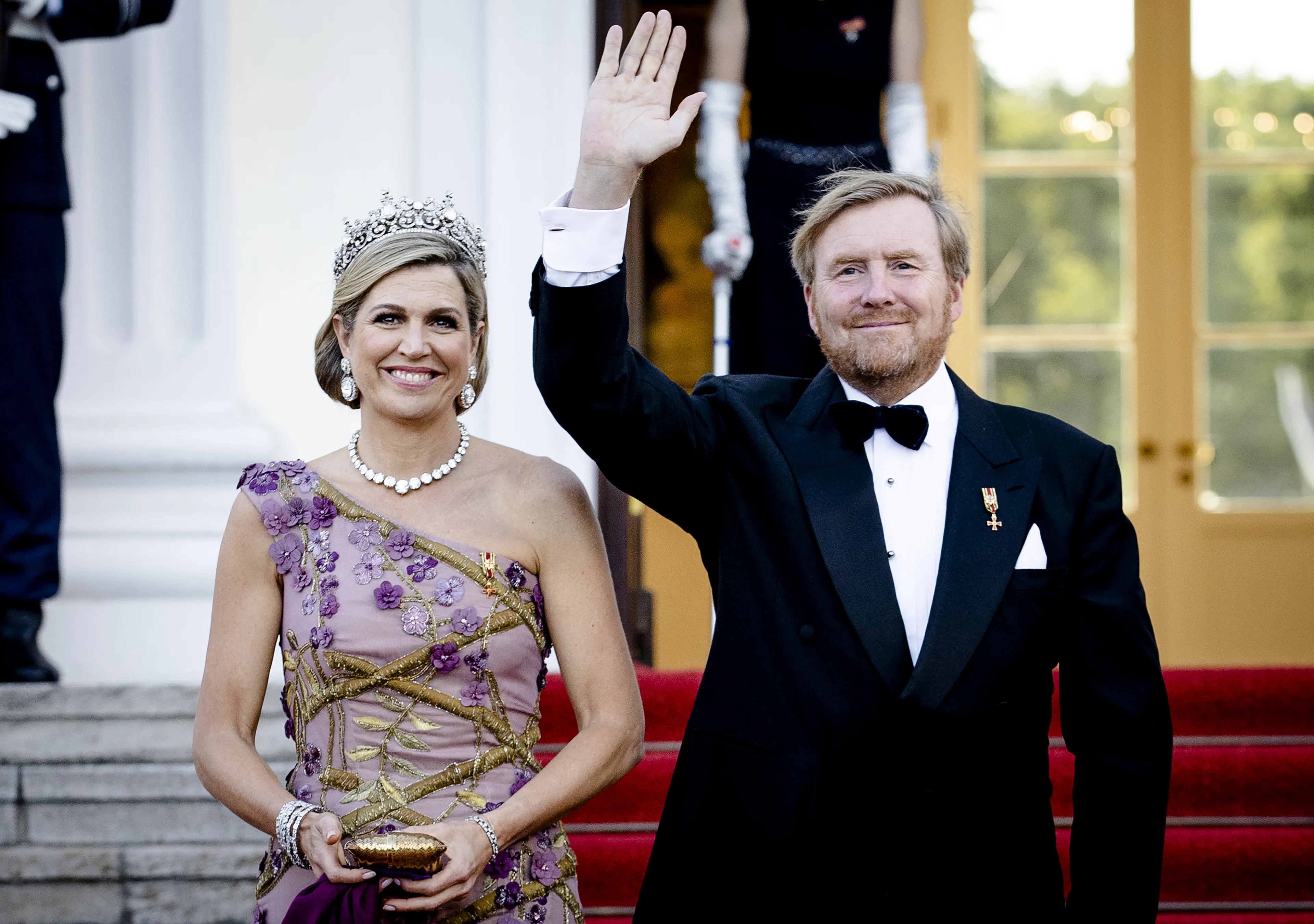 Koninklijk paar op een statig wit gebouw: koning Willem-Alexander en koningin Máxima staan op een met rood tapijt beklede bordestrap bij de ingang, gekleed in formele avondkleding en poseert als een ceremoniële, officiële moment.