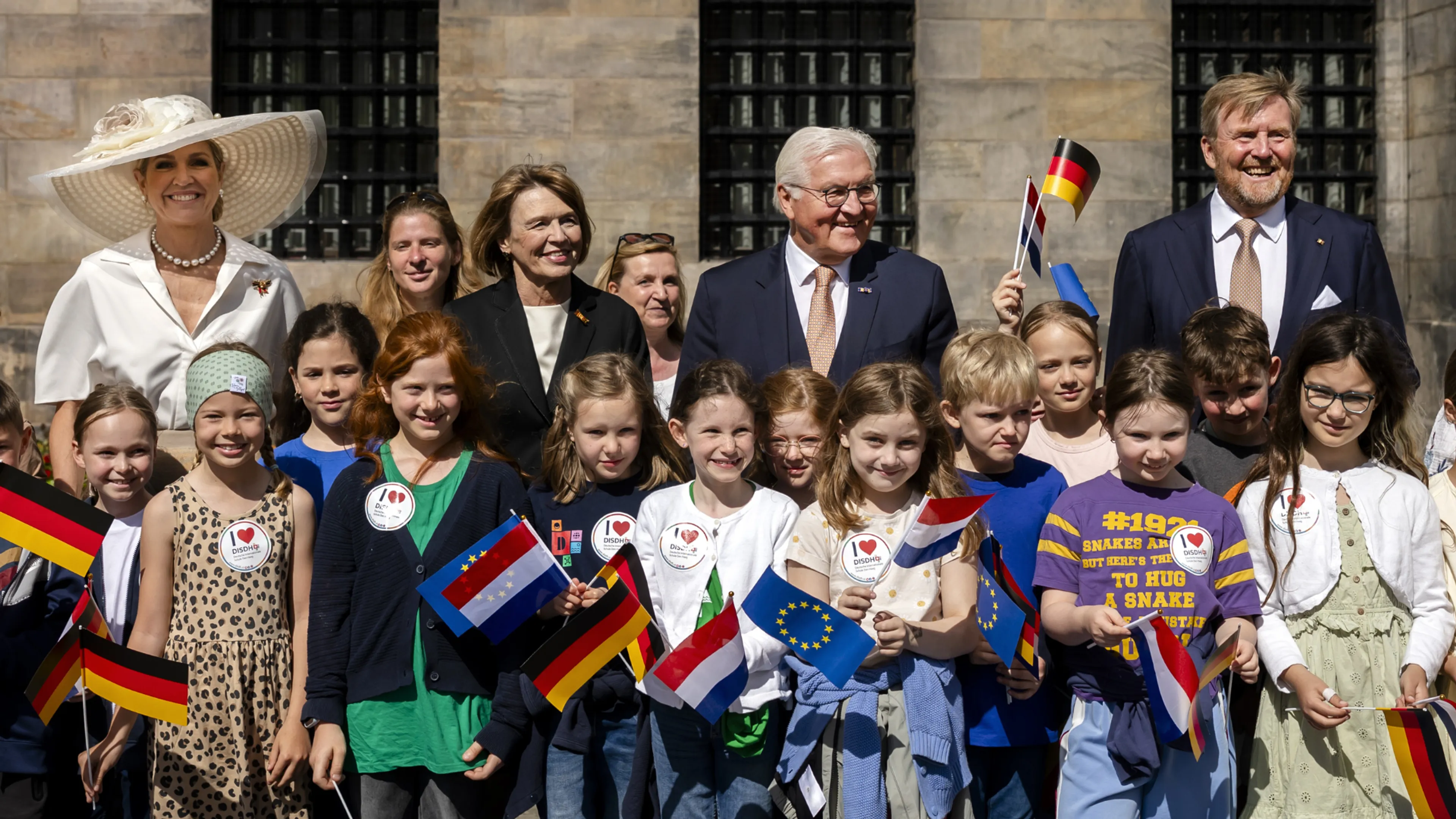 Koninklijk gezamenlijke schoolfoto (niet-officiële titel): Op een zonnige dag poseren ongeveer twintig basisschoolkinderen voor een monumentaal stenen gebouw met hoge, donkergeruite ramen, terwijl volwassenen in formele kleding achter hen staan; over de groep verspreid houden kinderen kleine vlaggen omhoog van Duitsland, Nederland en de Europese Unie, wat het internationale karakter van de gelegenheid benadrukt.